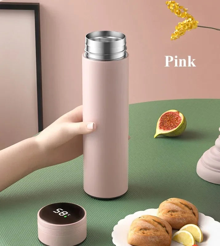 Pink thermos held by a hand on a green surface with figs and bread